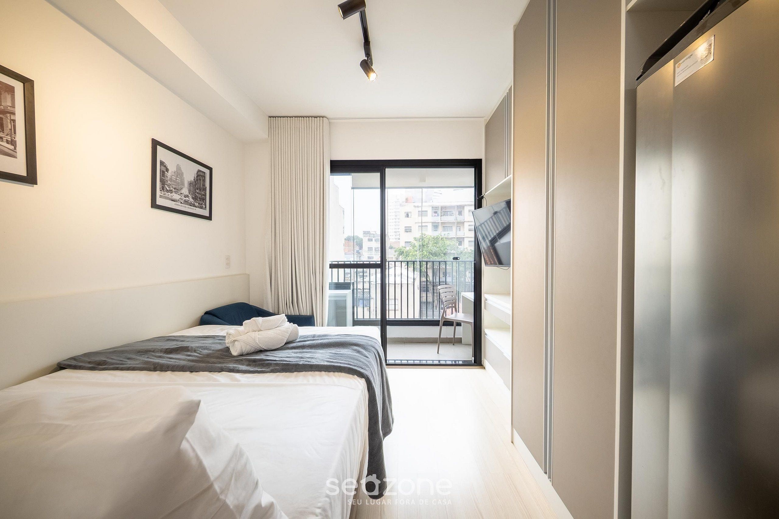 A modern hotel room with a bed, a balcony with a chair, and a city view.