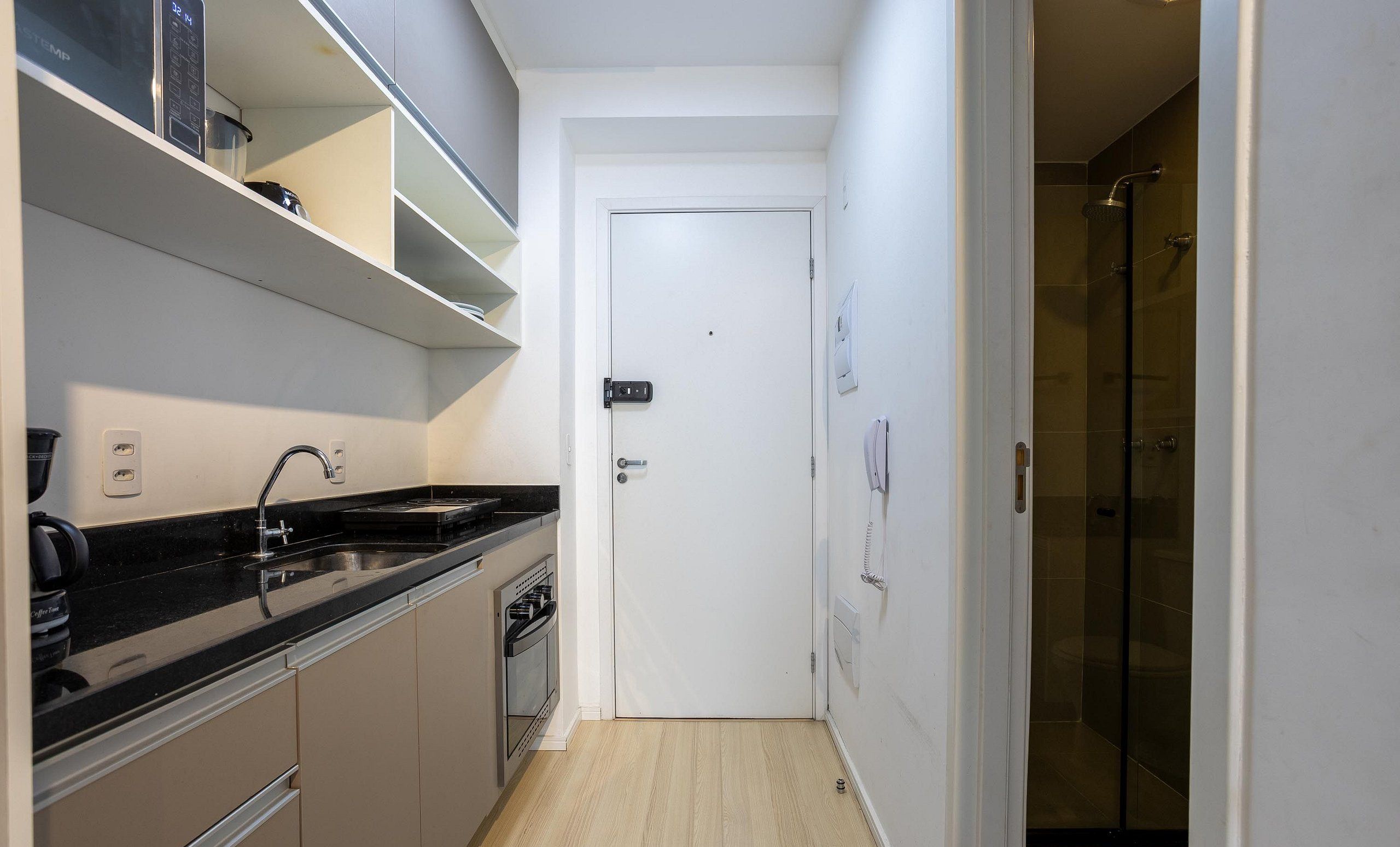 A narrow kitchen with a built-in oven and a coffee machine, a white door, and a shower area.
