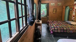 A wooden cabin room with a large window, a bed with a colorful quilt, and a small table with a lamp. - Chácara dos Cheiros: A cozy corner to relax. (Atibaia)
