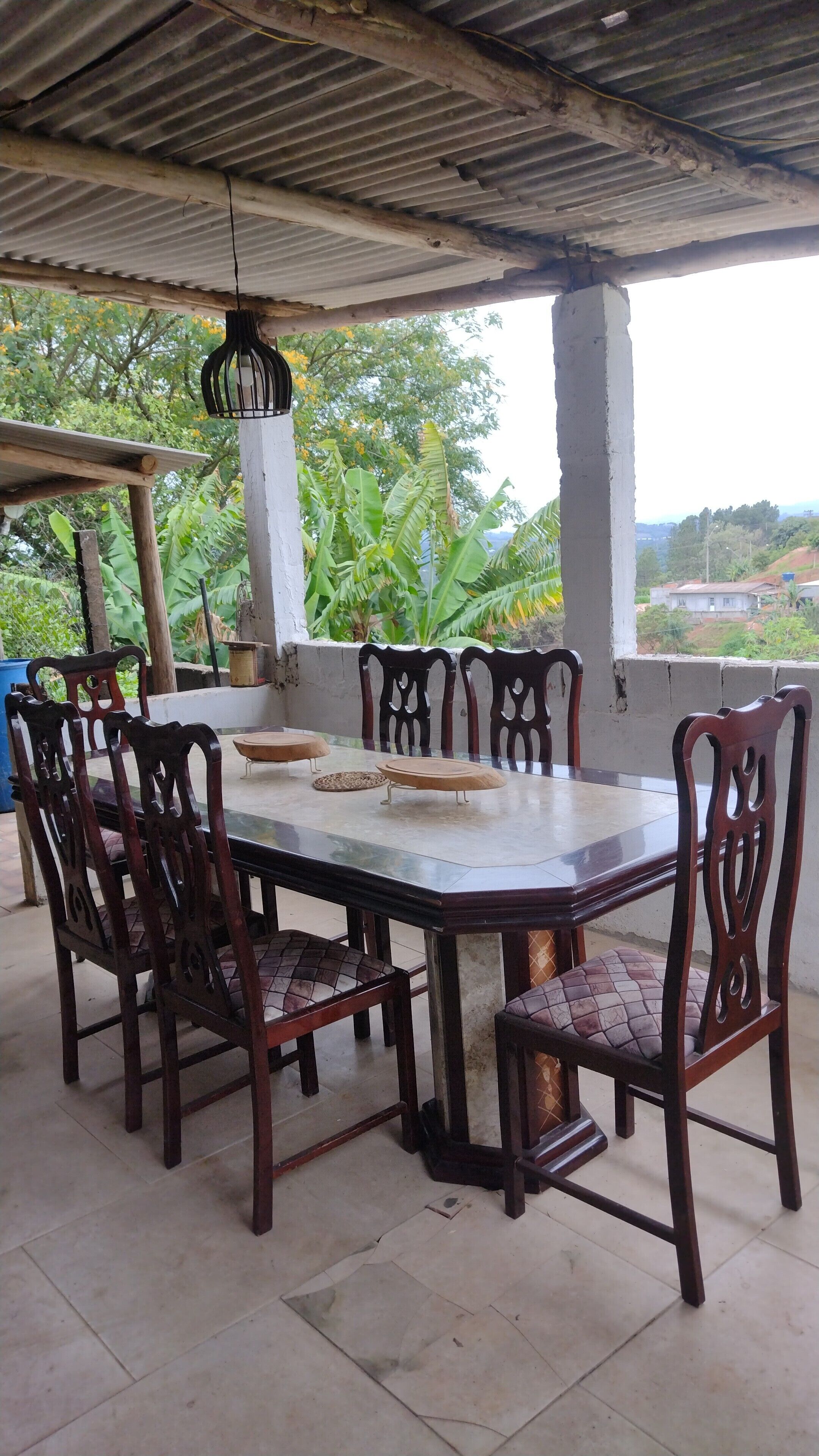 A covered outdoor dining area with a large table and six chairs, surrounded by greenery and a view of the landscape.