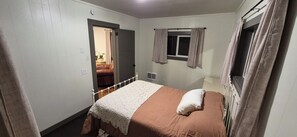 Una habitació amb un llit individual, una finestra amb cortines, una porta que condueix a una altra habitació i una taula petita amb una làmpada.