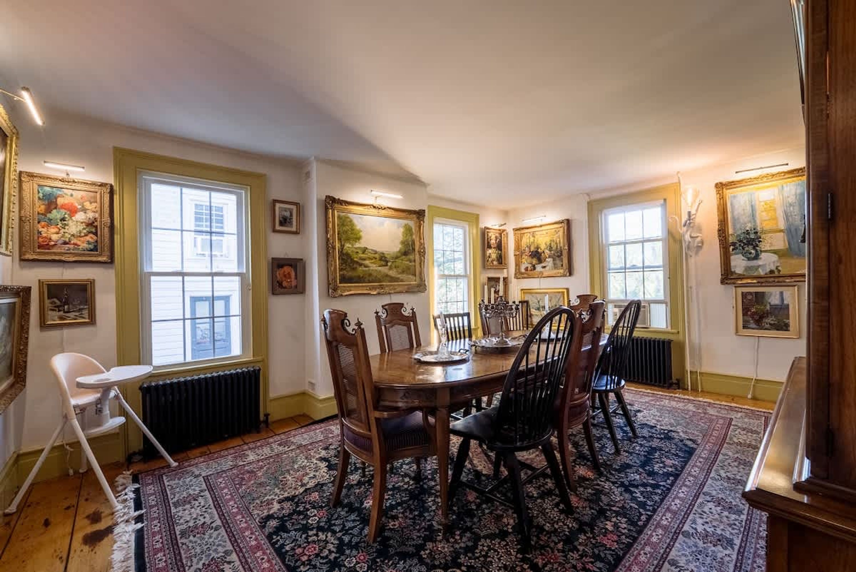Een eetruimte met een houten tafel, stoelen, een kamerkleed met dessin en kunstwerken aan de wanden.