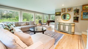 A spacious living room with a large sectional sofa, a round coffee table, and a piano. There are large windows offering a view of the outdoors. - Private Luxurious Canal Estate (Princeton)