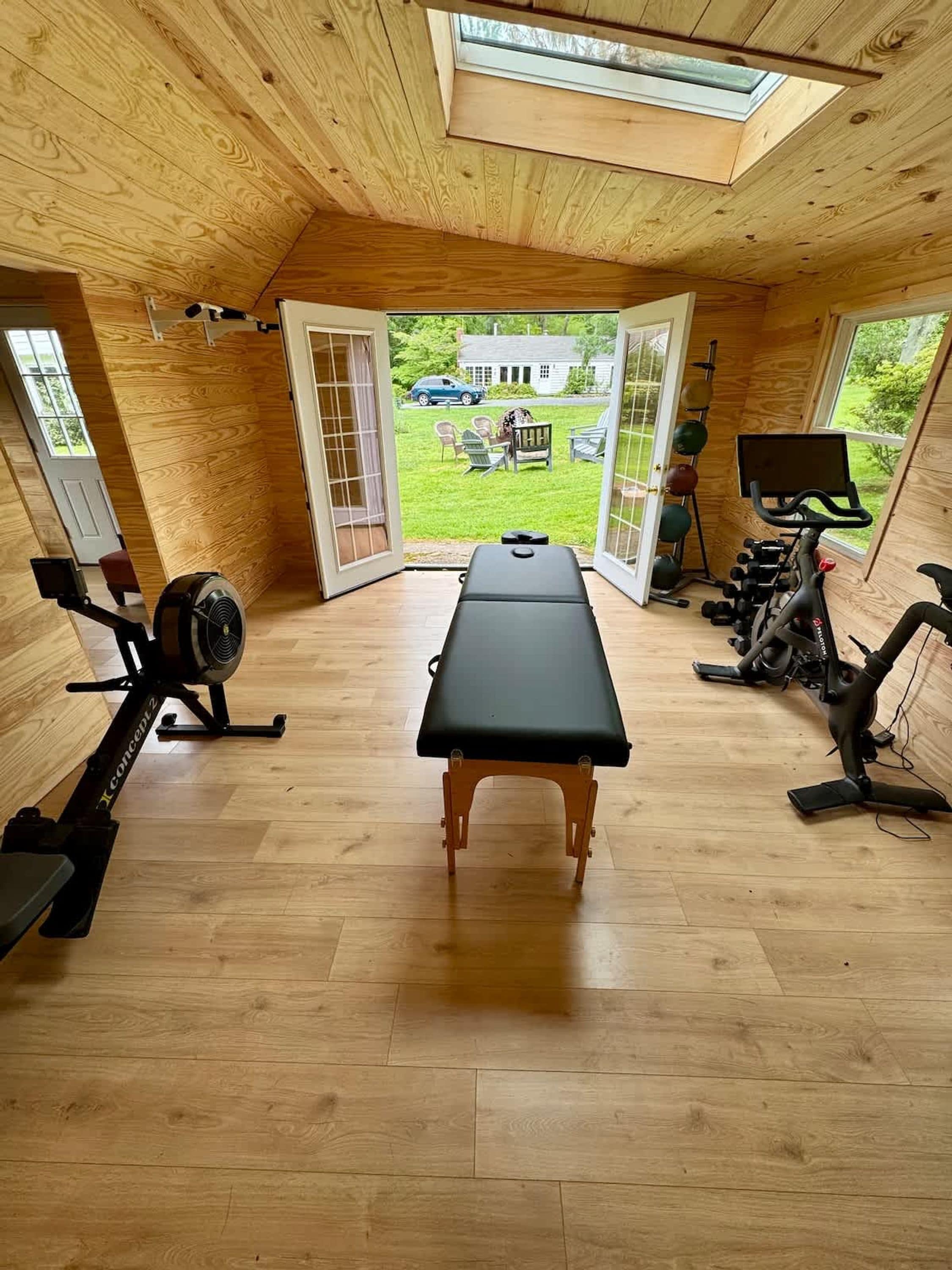 A spacious room with a wooden floor, a massage table, exercise equipment, and large windows offering a view of a house and lawn.