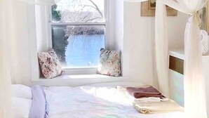 Een slaapkamer met een hemelbed, uitzicht op bomen door het raam, en een kamerkleed met een patroon.