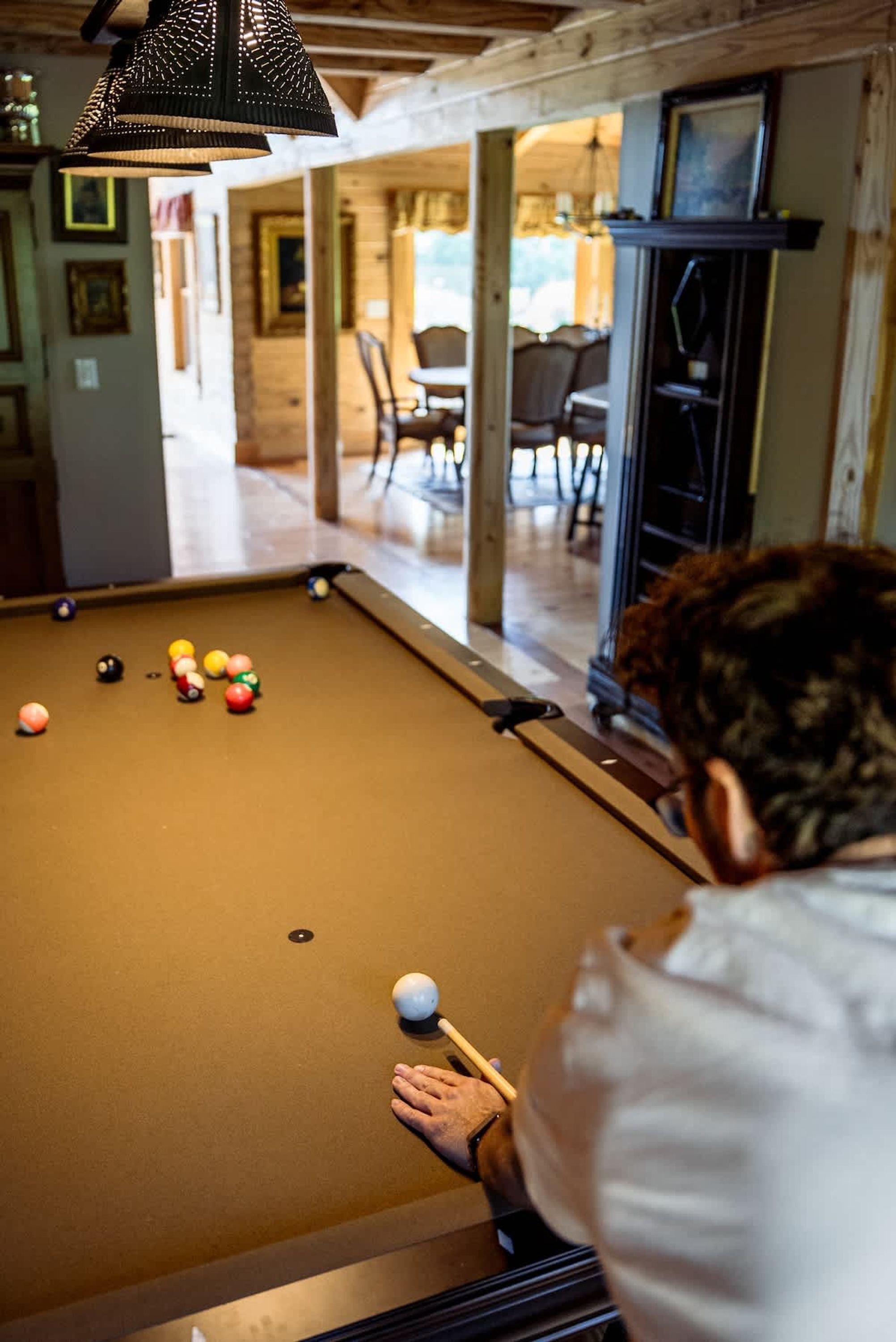 Una mesa de billar con bolas, una persona jugando y una habitación con vigas de madera y una lámpara colgante.