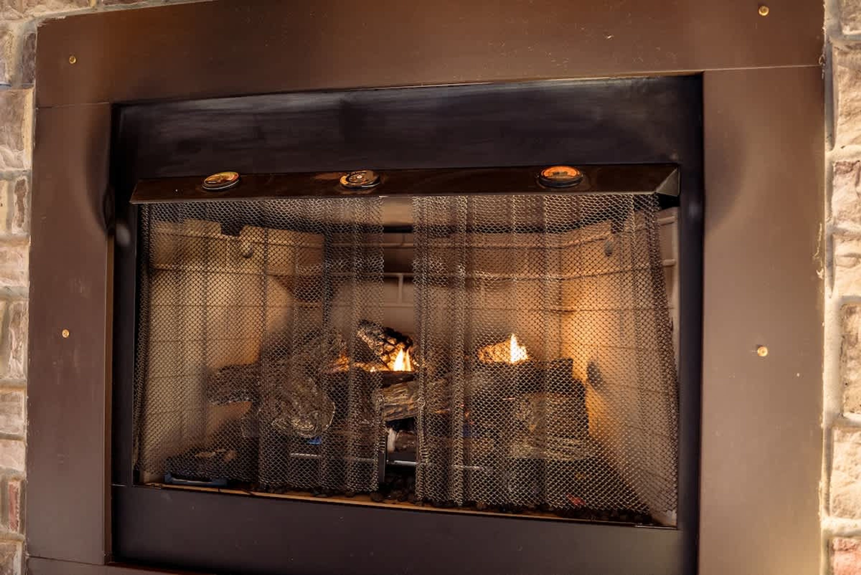 A fireplace with a metal screen, logs, and a brick wall.