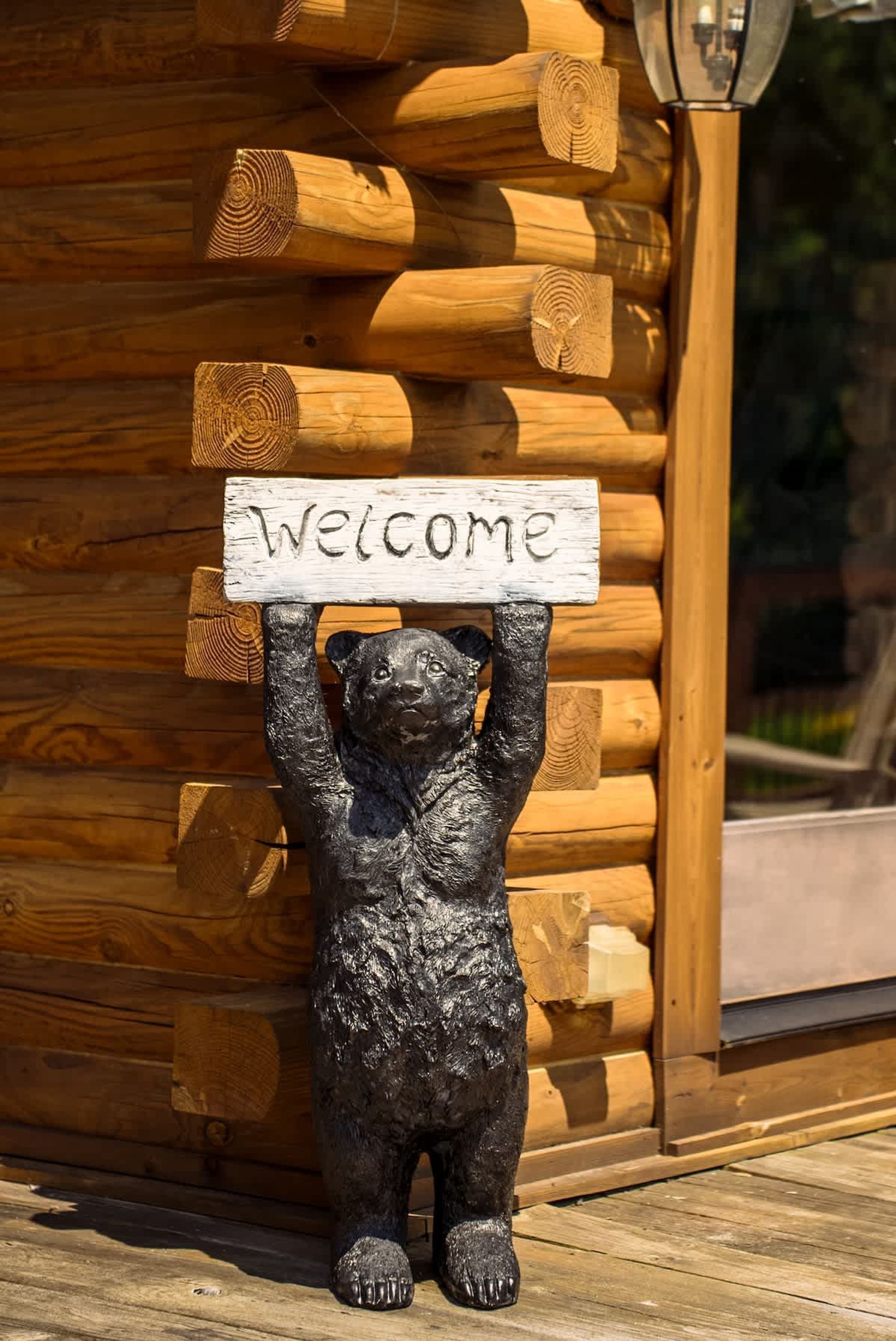 A bear holding a 'Welcome' sign in front of a log cabin.