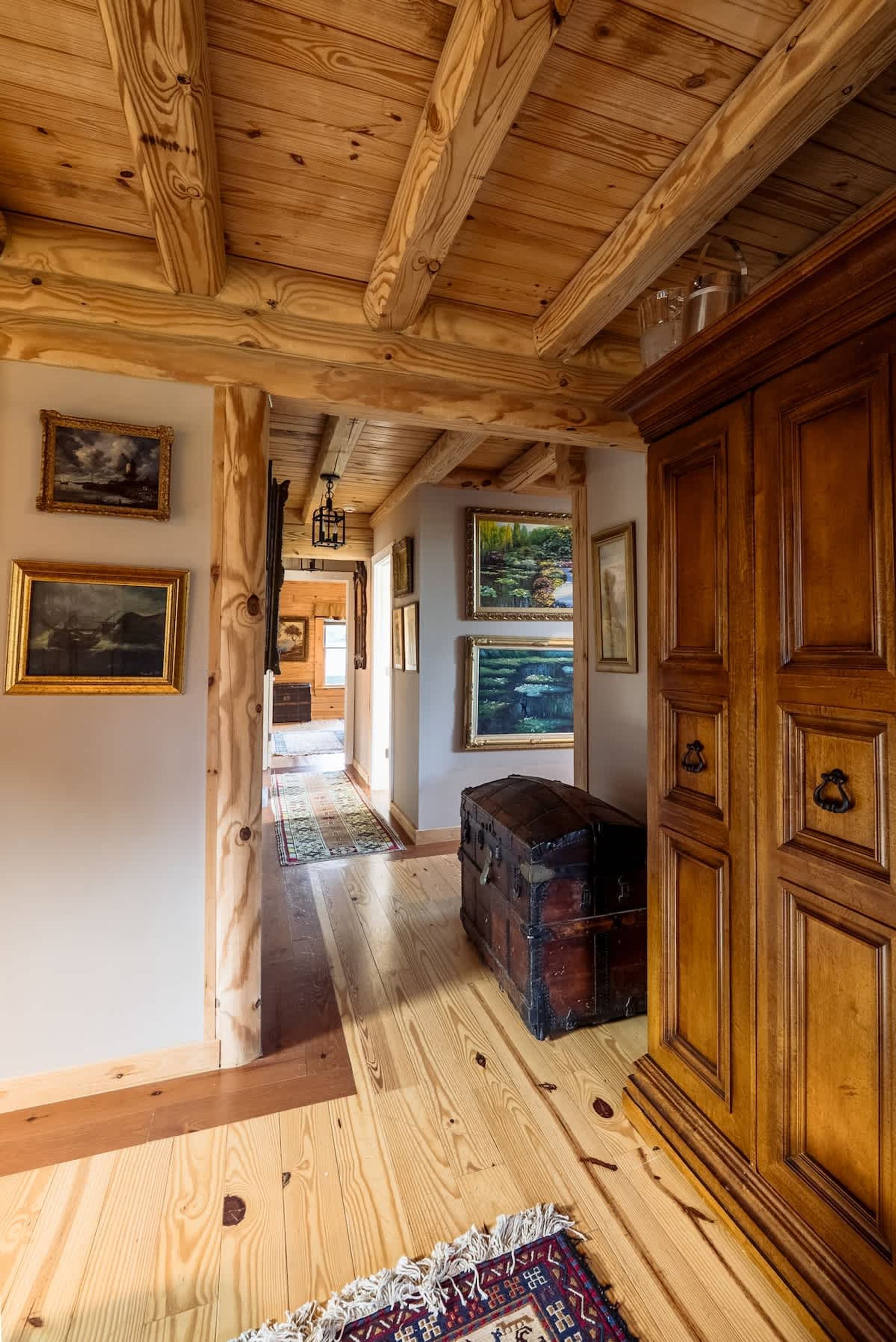 Interior em madeira com um corredor que leva a um quarto, apresentando vigas de madeira, um tapete e um armário de madeira grande.