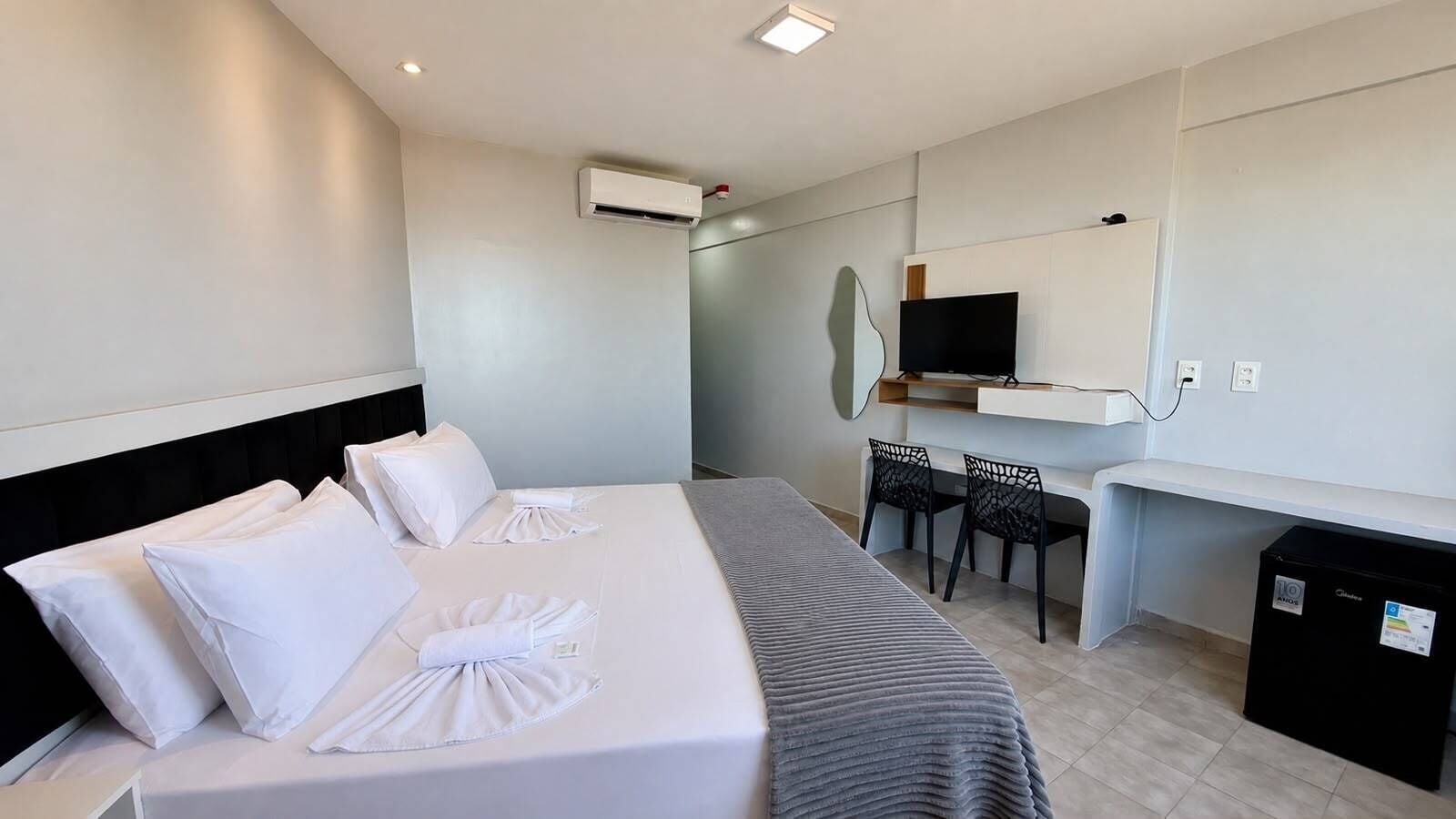 A hotel room with a bed, a desk, two chairs, a TV, and an air conditioning unit.