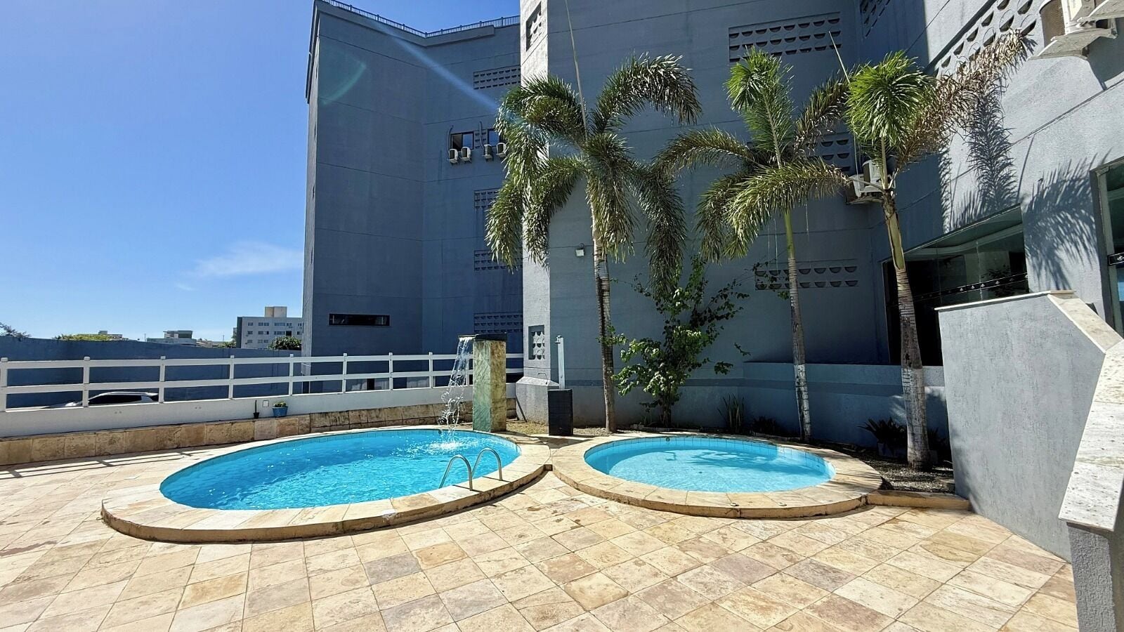 A rooftop pool with two circular pools, surrounded by palm trees and a white fence.