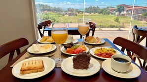 A table set with breakfast items including toast, eggs, fruit, and a slice of cake, with a view of a garden and buildings in the background. - Atlas Beach Hotel (Fortaleza)