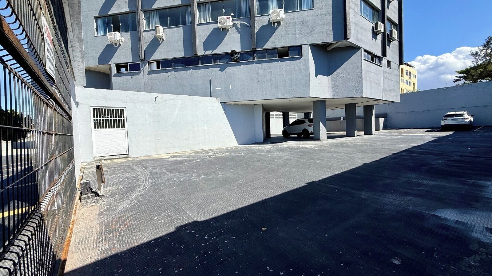 Un espace de stationnement avec une voiture blanche, un immeuble à plusieurs étages et des unités de climatisation à l’extérieur.