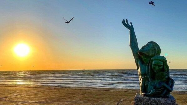 Una estatua de una mujer con la mano levantada, dominando una playa al atardecer.