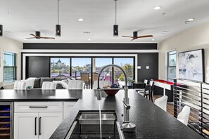 Una cocina moderna con una isla central, encimeras negras y electrodomésticos de acero inoxidable. Cuenta con ventiladores de techo, ventanas amplias con vistas a la ciudad y un comedor con sillas blancas.