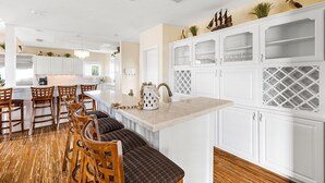 A modern kitchen with white cabinetry, a central island with seating, and a wooden dining area. - Lux West End Retreat |Fire Pit, 210 Steps to Beach (Galveston)
