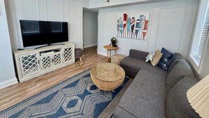 A living room with a flat-screen TV, a grey sofa, a round wicker ottoman, and a blue patterned rug. - 7 mins walk to Boardwalk & Beach! AC & Beach Tags! (Ventnor City)