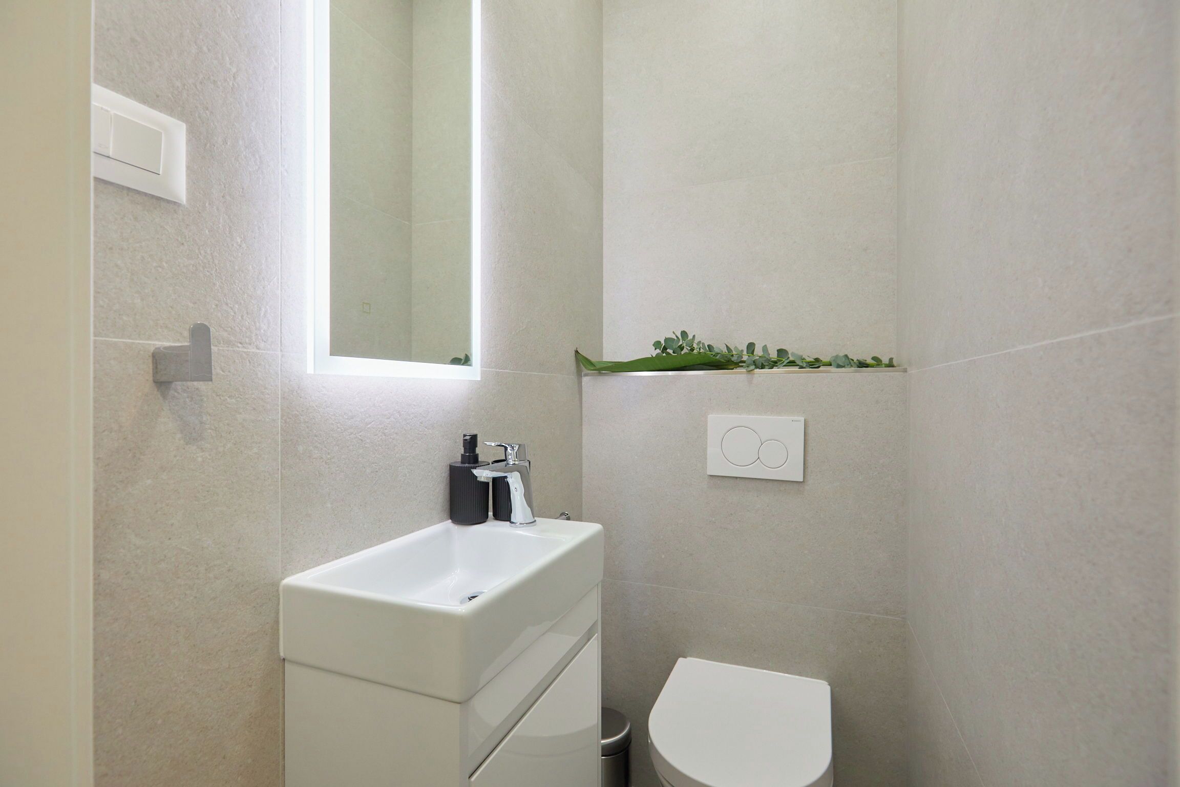 A bathroom with a white sink, a mirror, a toilet, and a small shelf with greenery.