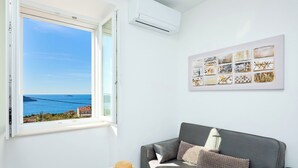 A room with a grey sofa, a white side table with a straw hat, a wall-mounted air conditioner, and a framed artwork. - Hedera Estate, House Hedera III (Dubrovnik)