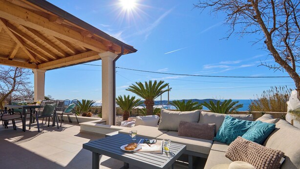 An outdoor patio with a wooden pergola, a seating area with a sofa and cushions, a table with a plate of food, and a view of the sea. - Hedera Estate, House Hedera III (Dubrovnik)