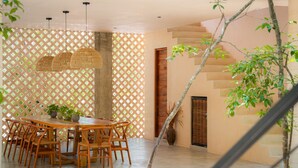 Un comedor con una mesa y sillas de madera, una lámpara colgante tejida y un separador de celosía con plantas.