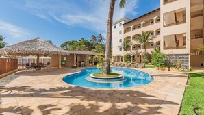 A swimming pool surrounded by a patio with lounge chairs, a thatched-roof structure, and a multi-story building with balconies. - Cumbuco Residence #309 - 2 Bedrooms by Carpediem (Caucaia)