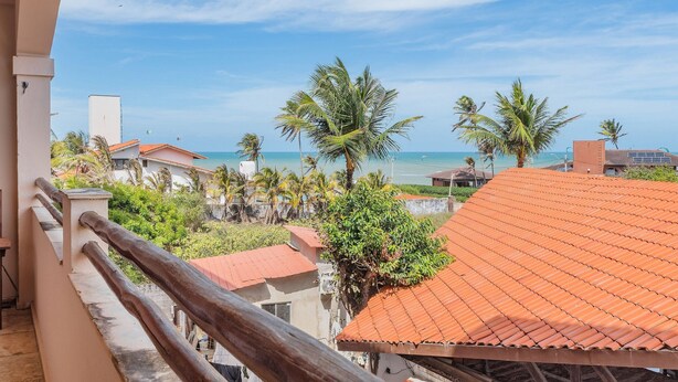 A balcony with a view of the sea, palm trees, and buildings with red roofs. - Cumbuco Residence #309 - 2 Bedrooms by Carpediem (Caucaia)
