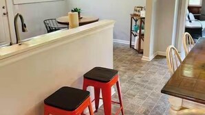 Una cocina con una isla central, dos taburetes rojos, una mesa de comedor con sillas y vistas al área de estar contigua.