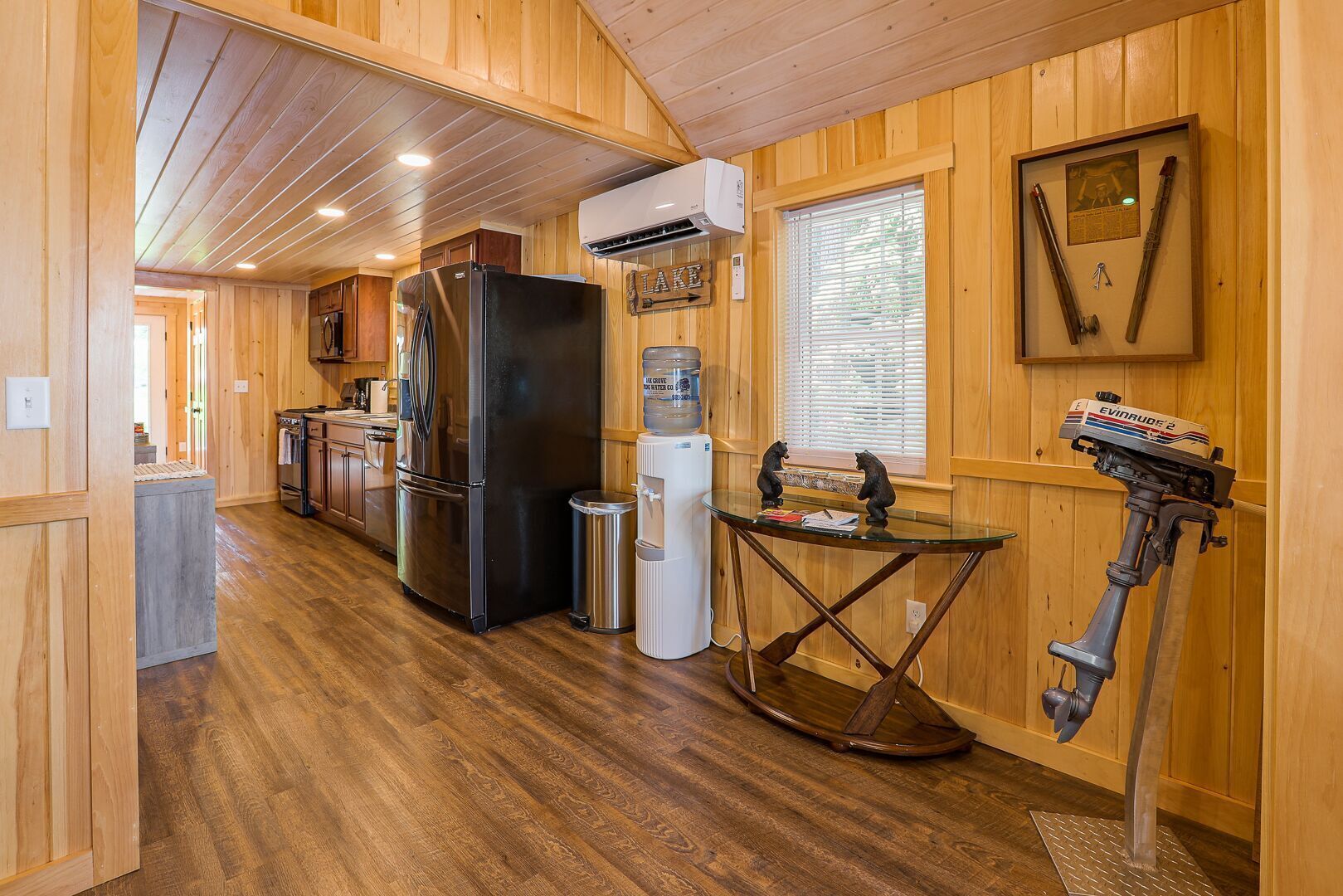 A compact kitchen with wooden walls, a refrigerator, a water dispenser, and a small table with a fishing rod.