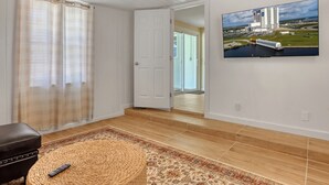 Una stanza con un pouf intrecciato, un telecomando sopra, un tappeto decorato, una TV a muro che mostra un'immagine di un centro spaziale e una porta che dà su un'altra stanza.