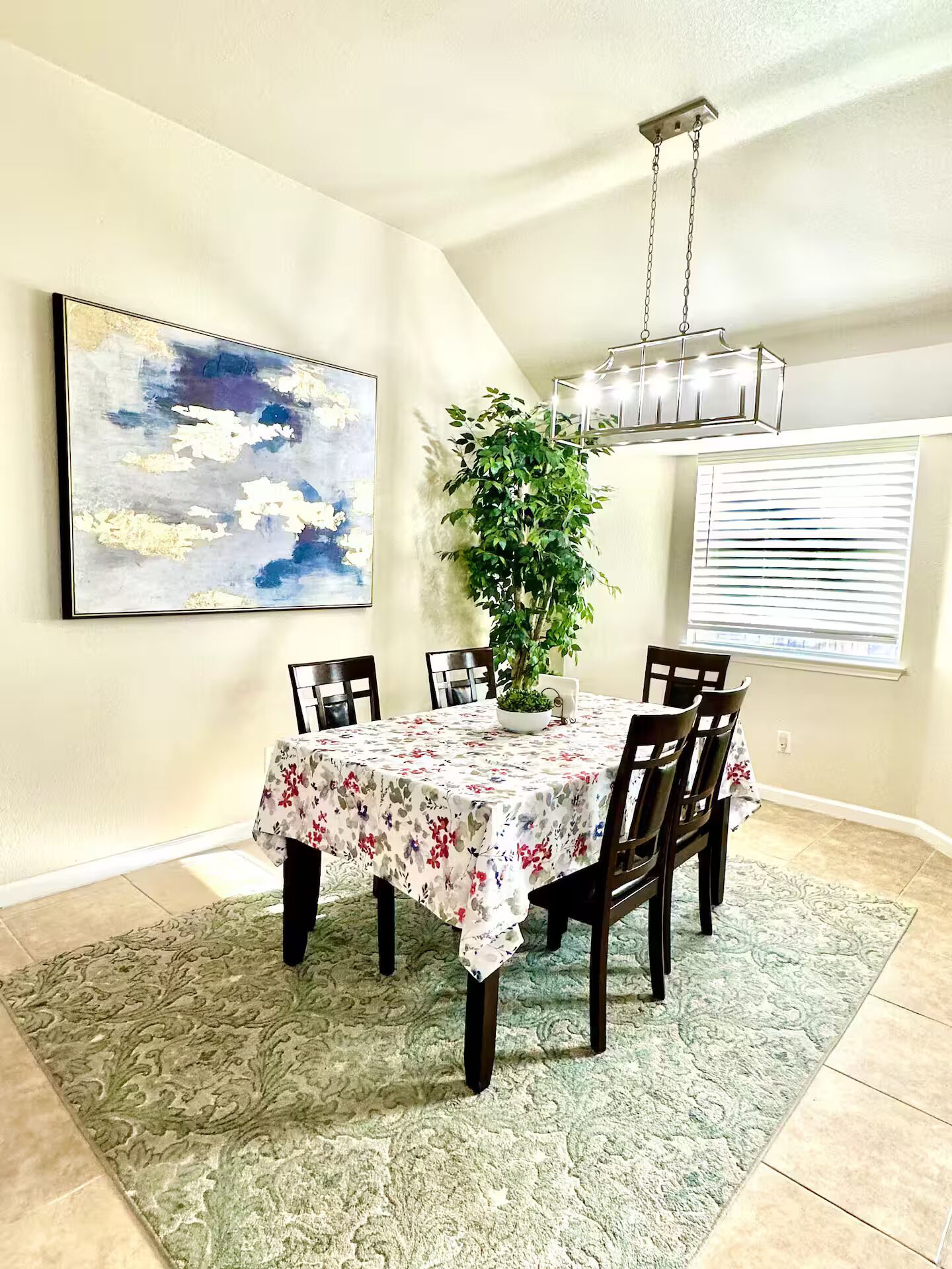 Un coin repas avec une table dressée pour quatre personnes, un luminaire suspendu, un grand tableau abstrait et une plante en pot.