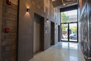 A hallway with a concrete wall, a fire alarm, and a door leading to an outdoor area with a view of a vehicle and trees. - Loft con Cama Matrimonial, Gym + Estacionamiento (Monterrey)