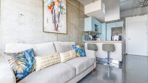 Un salon moderne comprenant un canapé, deux fauteuils, un coin cuisine et un grand tableau abstrait accroché au mur.