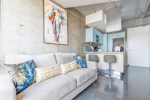 Un salon moderne comprenant un canapé, deux fauteuils, un coin cuisine et un grand tableau abstrait accroché au mur.