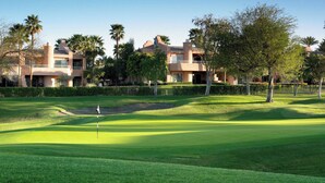 A golf course with a flagstick in the foreground, surrounded by well-maintained grass and a sand bunker. In the background, there are multi-story buildings with balconies, palm trees, and a clear sky. - Westin Mission Hills Resort Villas – 1BR Premium – March 7–14 2026 – Owner Week (Rancho Mirage)