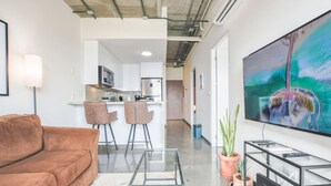 A modern living room with a brown sofa, a glass coffee table, a TV, and a kitchen in the background.