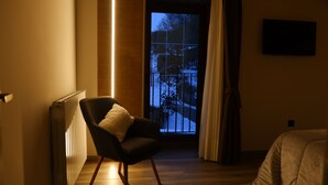 Een hotelkamer met een houten vloer, een zwarte fauteuil met een witte kussen, een radiator, een raam met uitzicht op bomen en een hek, en een flatscreen televisie aan de muur.