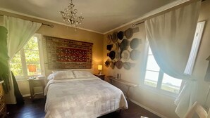 Ein Schlafzimmer mit einem großen Bett, einer Kristallleuchter, einer Wand voller Hüte und einem Fenster mit Vorhängen.
