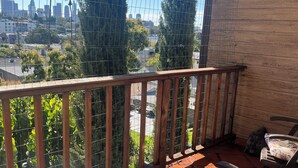 Ein hölzerner Balkon mit Metallgeländer, ein Stuhl mit gemustertem Kissen und Blick auf eine Stadtansicht durch ein Drahtgitter.