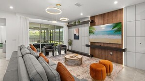 Una sala de estar moderna con un televisor grande, un sofá gris, otomanas naranjas y una mesa de centro redonda.