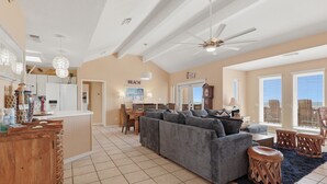 A spacious living area with a large gray sofa, a wooden coffee table, and a dining table with chairs. There is a kitchen area with a refrigerator and a wooden cabinet. The room has a high ceiling with a ceiling fan and a large window with blinds. - Casa Bonita Surfside (Freeport)