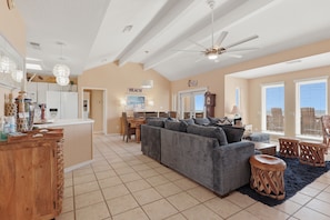 A spacious living area with a large gray sofa, a wooden coffee table, and a dining table with chairs. There is a kitchen area with a refrigerator and a wooden cabinet. The room has a high ceiling with a ceiling fan and a large window with blinds. - Casa Bonita Surfside (Freeport)