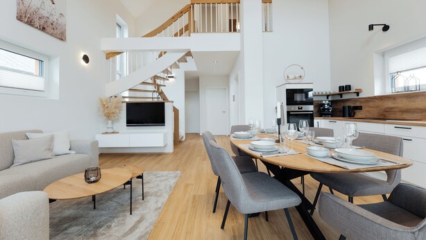 Ein geräumiger Wohnbereich mit Sofa, Couchtisch und Esstisch für sechs Personen. Eine Treppe führt zu einer oberen Ebene.