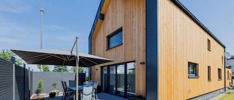 Ein modernes Haus mit Holzfassade, einer Terrasse und Gartenmöbeln sowie einem großen Fenster.