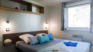 A neatly made bed with pillows and a book, a shelf with decorative items, and a window with curtains. - Palmyre - Confort 1 - 3p (1) (Les Mathes)