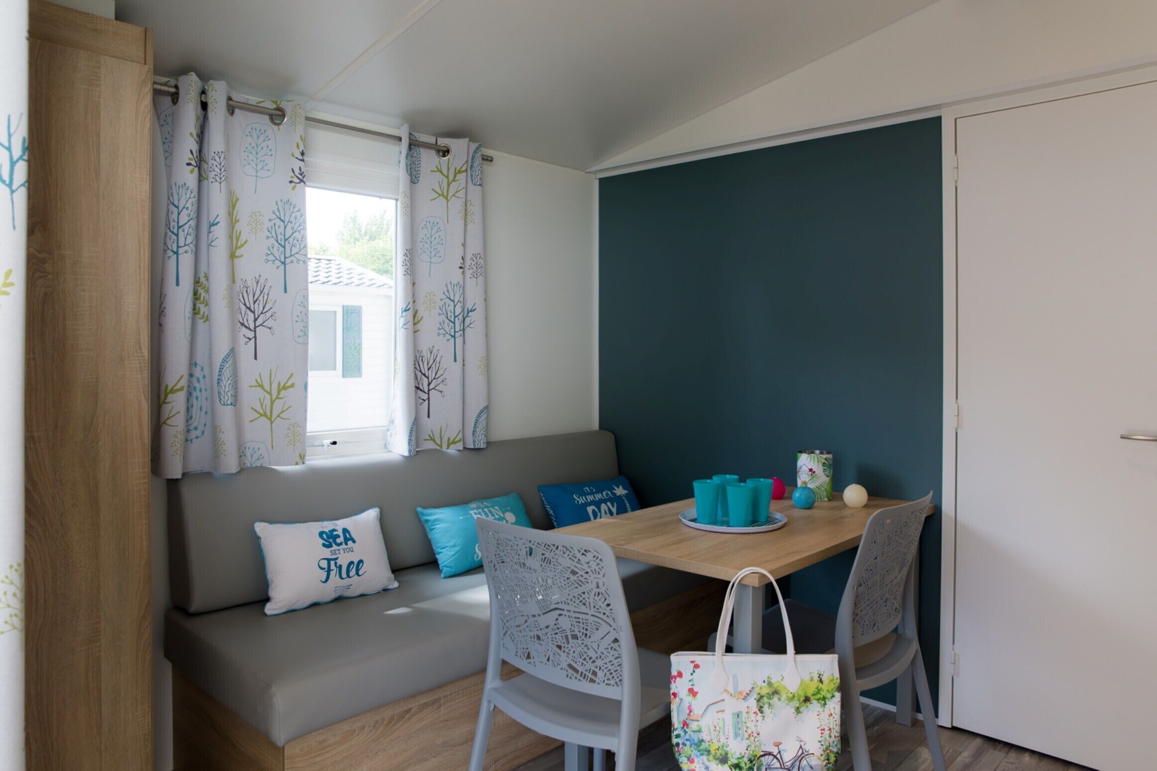 Um quarto acolhedor com uma mesa de madeira, um sofá cinza com almofadas azuis e uma janela com cortinas estampadas com motivos de árvores.