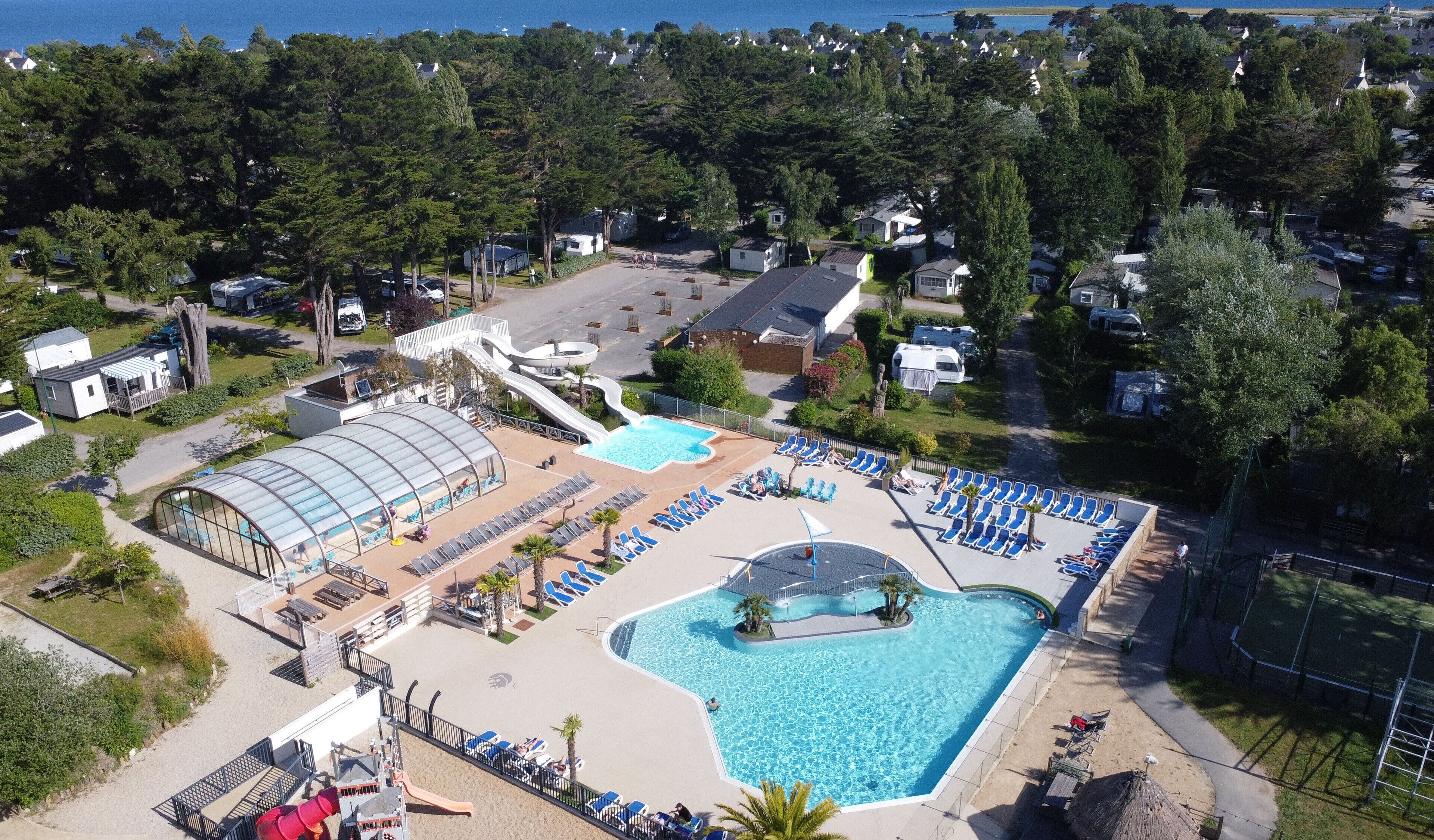 An aerial view of a resort with a large swimming pool, sun loungers, a covered area, and a small slide.