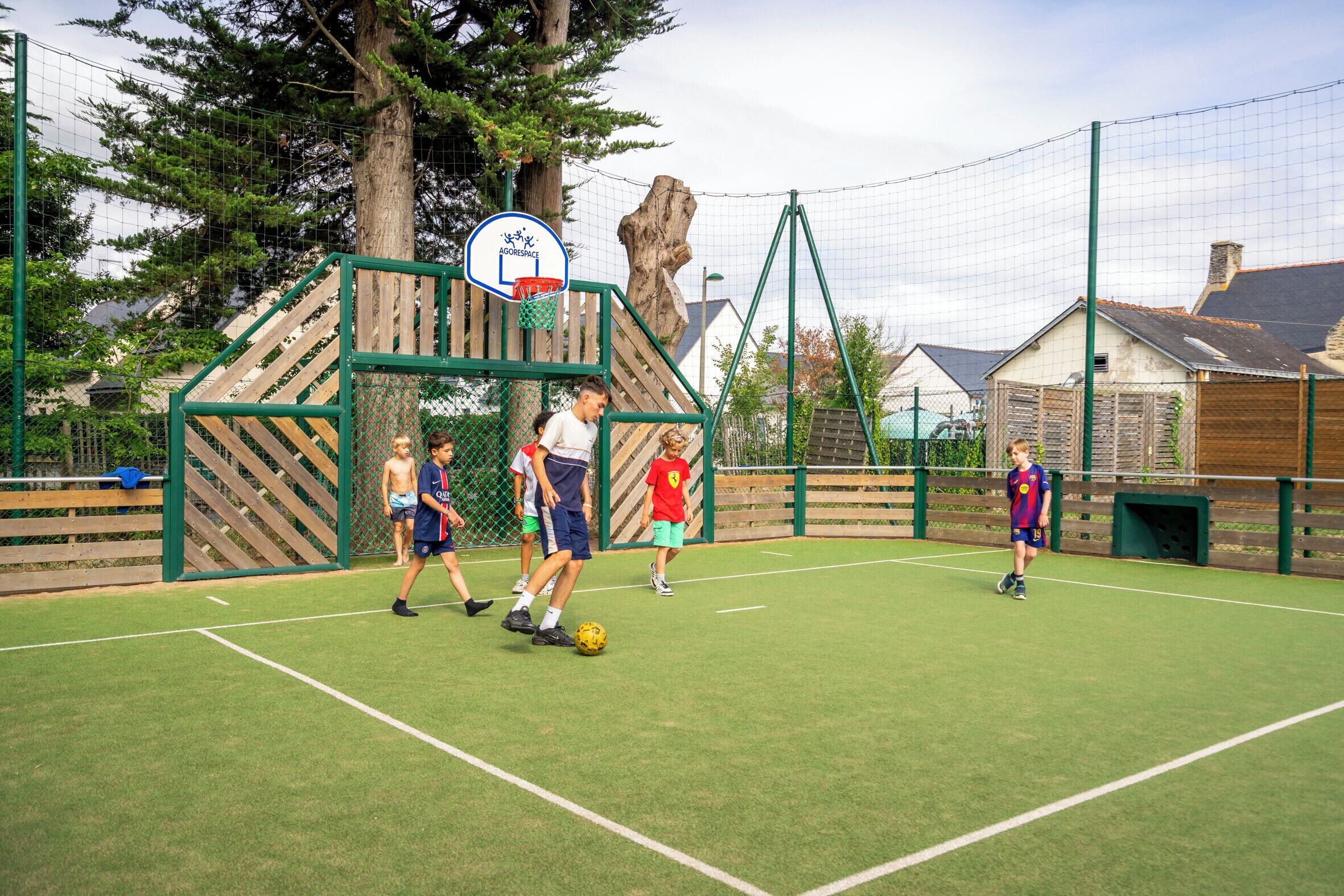 Ein Sportplatz mit einem Basketballkorb, umgeben von einem Zaun und Bäumen.