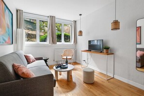 Een moderne woonkamer met een grijze bank, een ronde salontafel, een rieten fauteuil, een televisie op een houten dressoir en een groot raam met gordijnen.