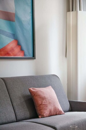 Een grijze bank met één roze kussen, een lijst met een abstract schilderij en een witte radiator.